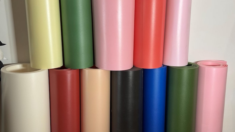 May include: A collection of rolled-up vinyl sheets in various colors, including yellow, green, pink, red, black, and blue. The rolls are stacked, showcasing the different hues and textures of the material.