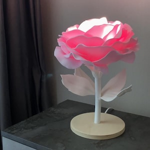 Puede incluir: Una lámpara en forma de rosa rosa con un tallo y hojas blancas. La lámpara está sobre una base de madera y está encendida.
