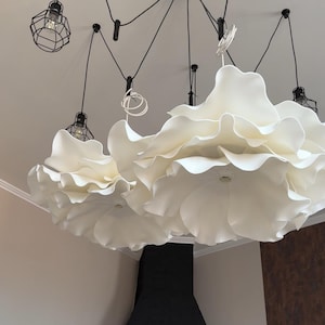 May include: A modern ceiling light fixture featuring large, white, flower-shaped shades. The fixture has multiple lights with black wire cages and black cords. The design is contemporary and elegant, suitable for various interior styles.