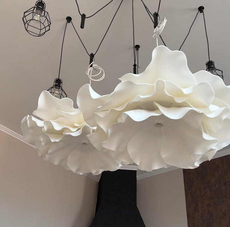 May include: A modern ceiling light fixture featuring large, white, flower-shaped shades. The fixture has multiple lights with black wire cages and black cords. The design is contemporary and elegant, suitable for various interior styles.