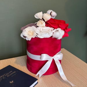 May include: A round, red velvet box with a white ribbon and bow, filled with white, cream, and red flowers, and cotton accents. A dark blue book with gold lettering sits to the left. The box is on a wooden surface.