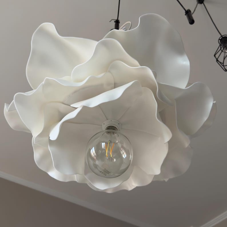 May include: A white, flower-shaped pendant light fixture. The light has multiple layers of large, petal-like shapes. A clear, round lightbulb is visible at the center. The fixture hangs from a black cord.
