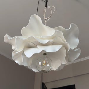 May include: A white, flower-shaped pendant light fixture. The light has multiple layers of petal-like shapes, with a clear, round lightbulb at the center. The fixture hangs from the ceiling by a black cord and white wire.
