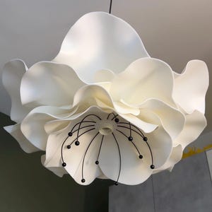 Puede incluir: Una lámpara colgante blanca con forma de flor grande y abierta, con pétalos superpuestos y ondulados. El centro presenta un casquillo rodeado de finos estambres de alambre negro curvado, cada uno con una pequeña cuenta negra redonda.