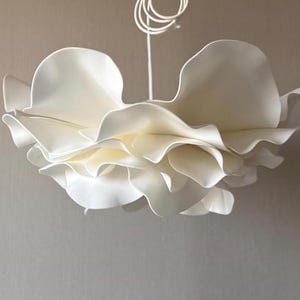 Può includere: Una lampada a sospensione bianca caratterizzata da molteplici strati di paralumi curvi e increspati a forma di petalo che creano una forma organica voluminosa. La lampada è sospesa da un sottile cavo bianco che si avvolge in alto.