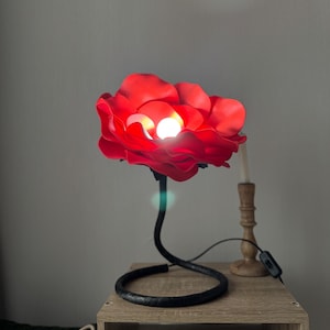 Può includere: Una lampada a forma di rosa rossa con una luce intensa al centro. La lampada ha uno stelo nero curvo e una base circolare. È appoggiata su un comodino di legno, accanto a un candeliere.