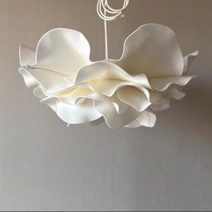 Puede incluir: Lámpara colgante blanca con forma de flor. La lámpara presenta múltiples capas de formas similares a pétalos, creando un diseño suave y fluido. Está suspendida de un cable blanco.