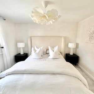 Puede incluir: Un dormitorio con una cama blanca con un cabecero alto tapizado, dos mesitas de noche negras con lámparas blancas y una gran lámpara colgante blanca en forma de flor que cuelga sobre la cama, creando un espacio luminoso y neutro.