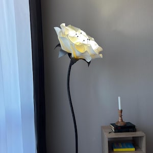Puede incluir: Una lámpara de pie única con forma de rosa blanca grande y abierta. La lámpara tiene un tallo negro y hojas de color gris oscuro. Los pétalos de la rosa están iluminados, creando un brillo suave. Una pequeña mesa auxiliar de madera con libros y una vela está al lado de la lámpara.