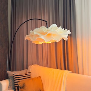 May include: A floor lamp with a thin, black, arched metal stand and a large, white, ruffled, cloud-like lampshade that emits a warm, golden glow. The lamp is positioned over a white sofa adorned with several decorative throw pillows and a blanket.