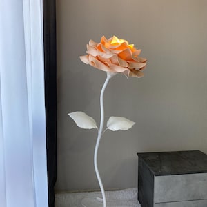 Op de afbeelding: Een unieke vloerlamp in de vorm van een gigantische roos. De lamp heeft een perzikroze bloemhoofd met een heldergeel centrum, ondersteund door een witte stengel en twee witte bladeren. De basis is een ronde houten schijf. De lamp is aangesloten.