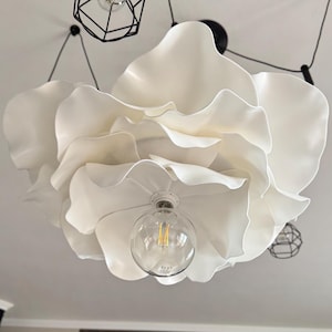 May include: A large, white, flower-shaped ceiling light fixture with multiple layered petals. The light has a clear, round bulb in the center, and is suspended from the ceiling by black cords. Additional black geometric light fixtures are also visible.