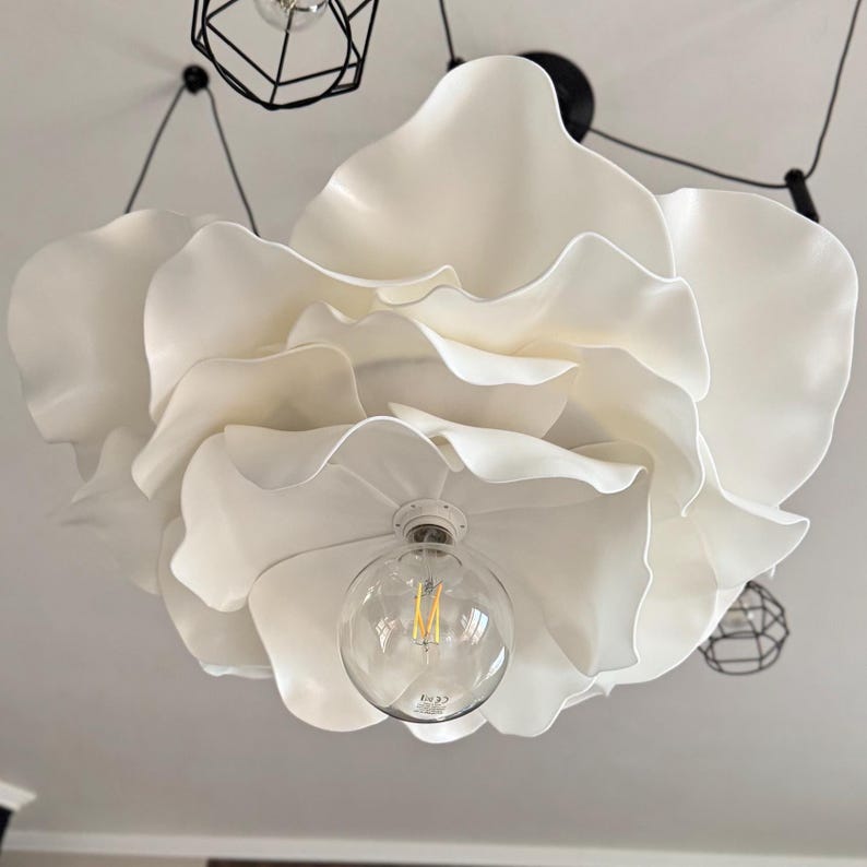 May include: A large, white, flower-shaped ceiling light fixture with multiple layered petals. The light has a clear, round bulb in the center, and is suspended from the ceiling by black cords. Additional black geometric light fixtures are also visible.