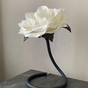 Può includere: Un grande fiore di carta bianco con stelo e base neri. Il fiore è su una superficie grigia testurizzata.