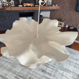 May include: A large, off-white, flower-shaped pendant light fixture. The light has multiple layers of petal-like shapes, creating a voluminous, sculptural design. The fixture hangs from a white cord, and is suspended from the ceiling.