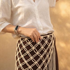 May include: A white button-down shirt paired with a dark brown skirt featuring an off-white macrame overlay. The macrame layer is constructed with a diamond-shaped knotted grid pattern and long, vertical fringe strands hanging from the bottom edge.
