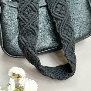 May include: Black handbag with a textured surface and a macrame strap. The strap features a woven design. White flowers are in the foreground.