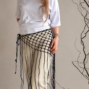 May include: A black crochet net hip wrap featuring a wide-mesh grid pattern and long, dangling fringe strands that extend downward. The wrap is secured at the side with thin, knotted ties, creating an open, layered look over light-colored trousers.