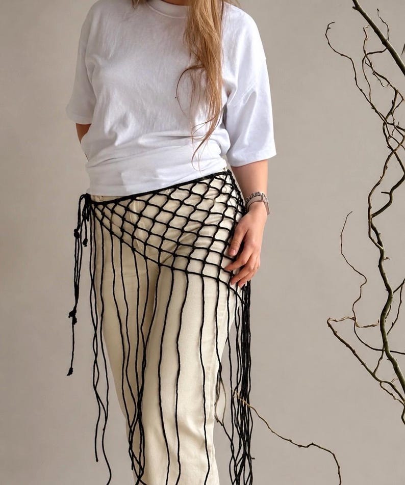 May include: A black crochet net hip wrap featuring a wide-mesh grid pattern and long, dangling fringe strands that extend downward. The wrap is secured at the side with thin, knotted ties, creating an open, layered look over light-colored trousers.