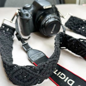 May include: A black Canon DSLR camera with a black macrame camera strap. The strap has a woven design and a red accent. The camera lens is black, and the strap is attached with metal clips.