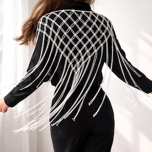 May include: A black long-sleeved shirt featuring a white macrame overlay on the back. The macrame design consists of a diamond-shaped knotted grid pattern that extends from the shoulders down to the mid-back, ending in long, loose fringe.