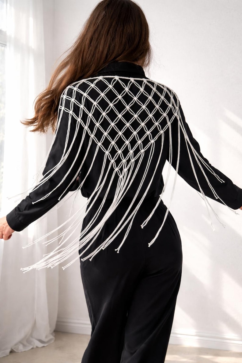 May include: A black long-sleeved shirt featuring a white macrame overlay on the back. The macrame design consists of a diamond-shaped knotted grid pattern that extends from the shoulders down to the mid-back, ending in long, loose fringe.