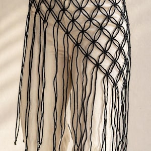 May include: A black macrame waist accessory featuring a knotted lattice grid pattern that forms a diamond shape. The piece includes long, dangling cord fringe that extends downward, with small knots tied at the ends of each individual cord strand.