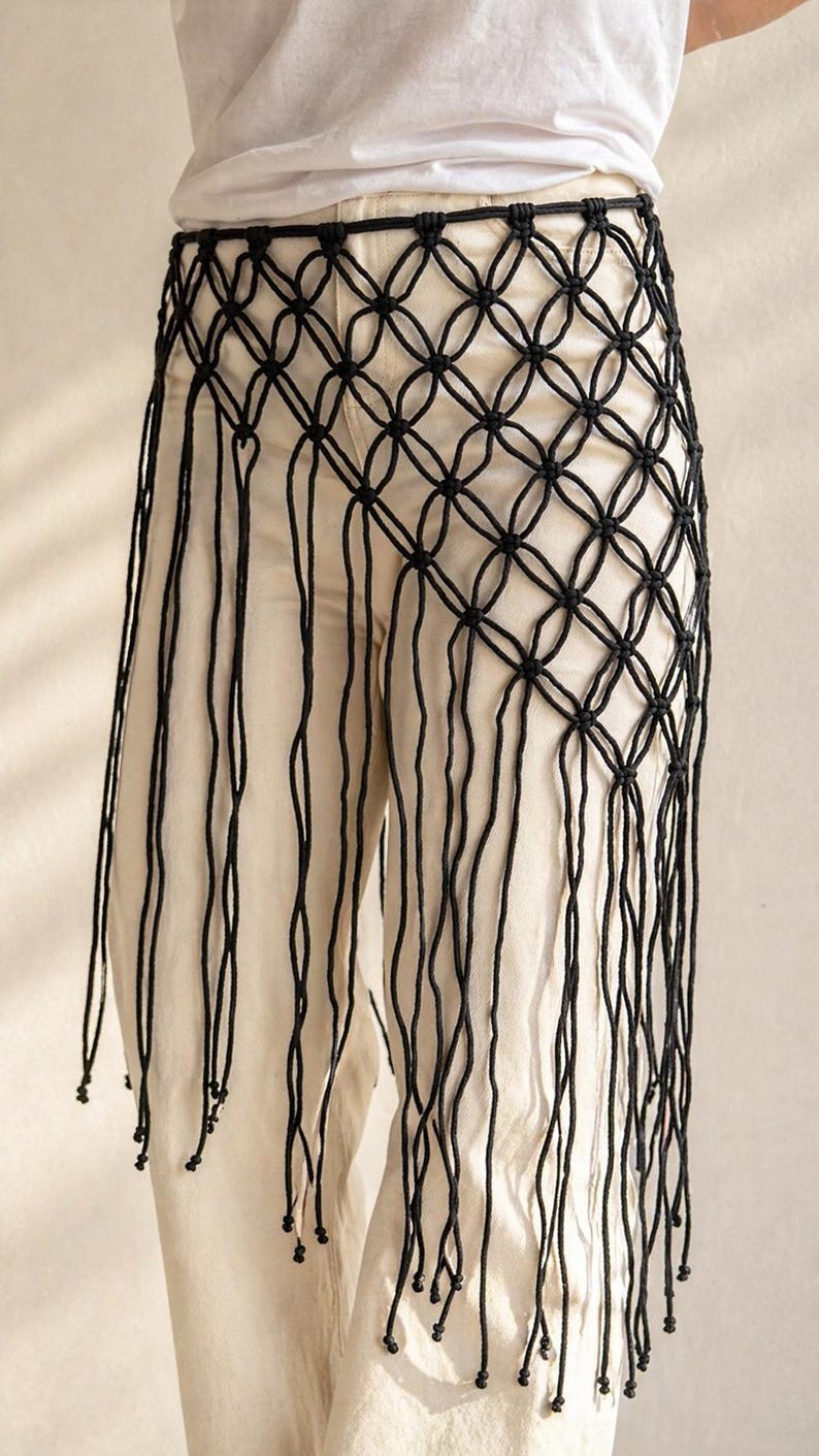 May include: A black macrame waist accessory featuring a knotted lattice grid pattern that forms a diamond shape. The piece includes long, dangling cord fringe that extends downward, with small knots tied at the ends of each individual cord strand.