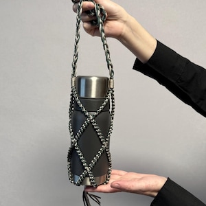 May include: A matte dark gray cylindrical metal bottle encased in a decorative, hand-knotted paracord net holder. The holder features a diamond-patterned weave in shades of forest green and silver, with a long braided carrying strap and tassel.
