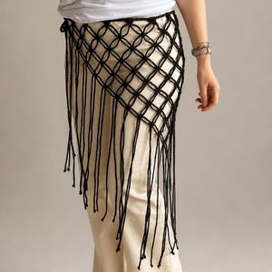 May include: A black macrame waist accessory featuring a knotted diamond grid pattern and long, dangling fringe strands. The piece is worn over cream-colored trousers and a white t-shirt, showcasing a structured, open-weave design.