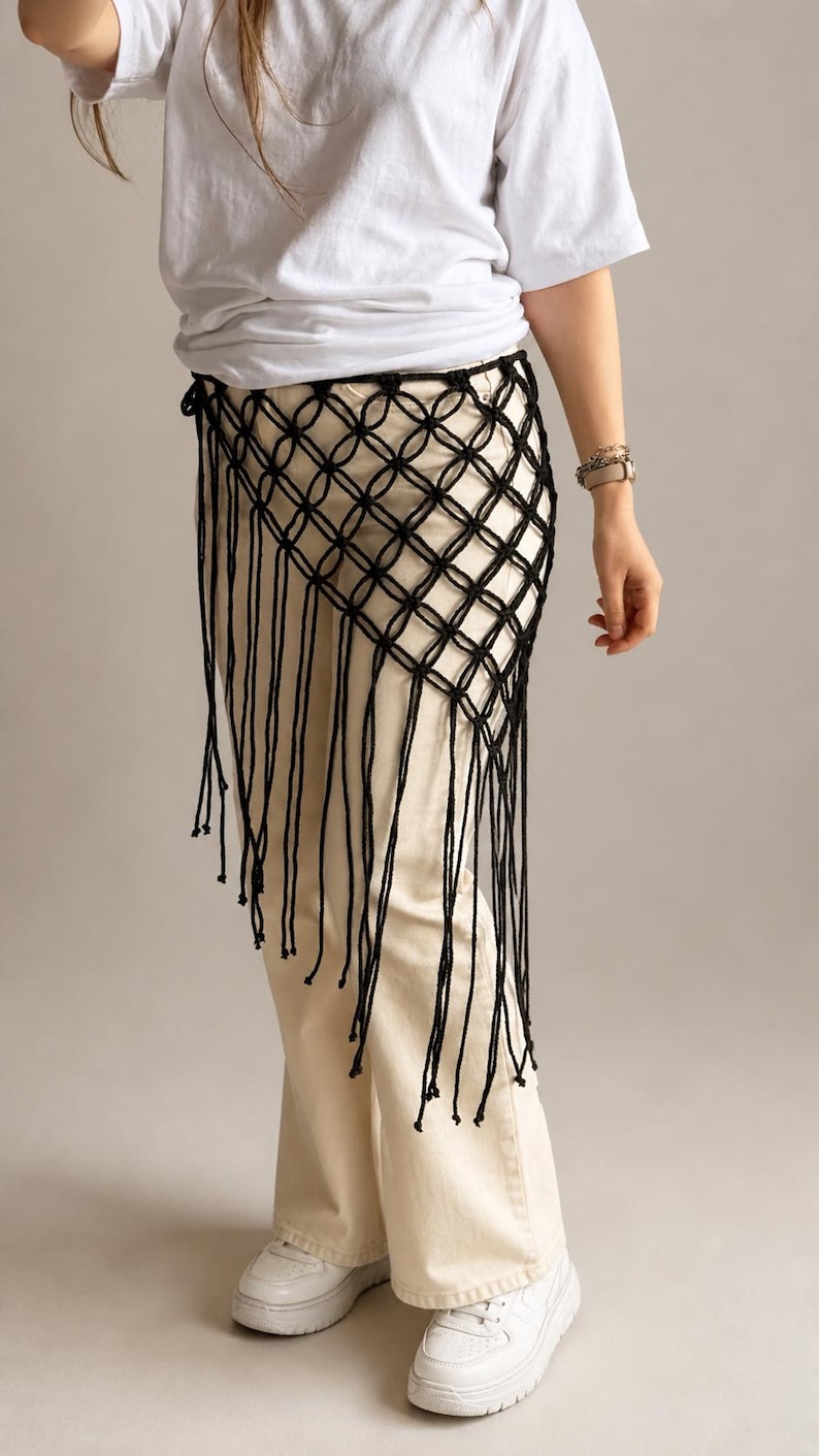 May include: A black macrame waist accessory featuring a knotted diamond grid pattern and long, dangling fringe strands. The piece is worn over cream-colored trousers and a white t-shirt, showcasing a structured, open-weave design.