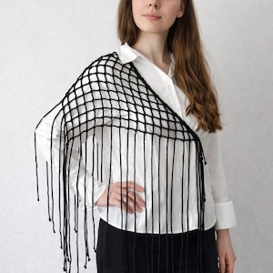 May include: A black crochet shawl featuring an open grid pattern and long, dangling fringe along the bottom edge. The accessory is draped over one shoulder, showcasing the knotted yarn construction and the uniform length of the hanging strands.