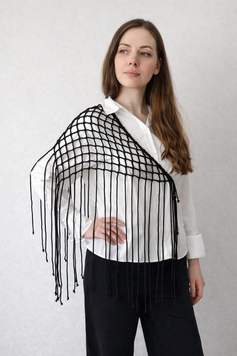 May include: A black crochet shawl featuring an open grid pattern and long, dangling fringe along the bottom edge. The accessory is draped over one shoulder, showcasing the knotted yarn construction and the uniform length of the hanging strands.