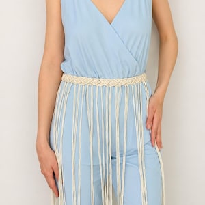 May include: A light blue sleeveless jumpsuit with a V-neckline and thin spaghetti straps, featuring a cream-colored macrame belt at the waist. The belt has a woven pattern and long, dangling fringe cords that extend down the front of the jumpsuit.
