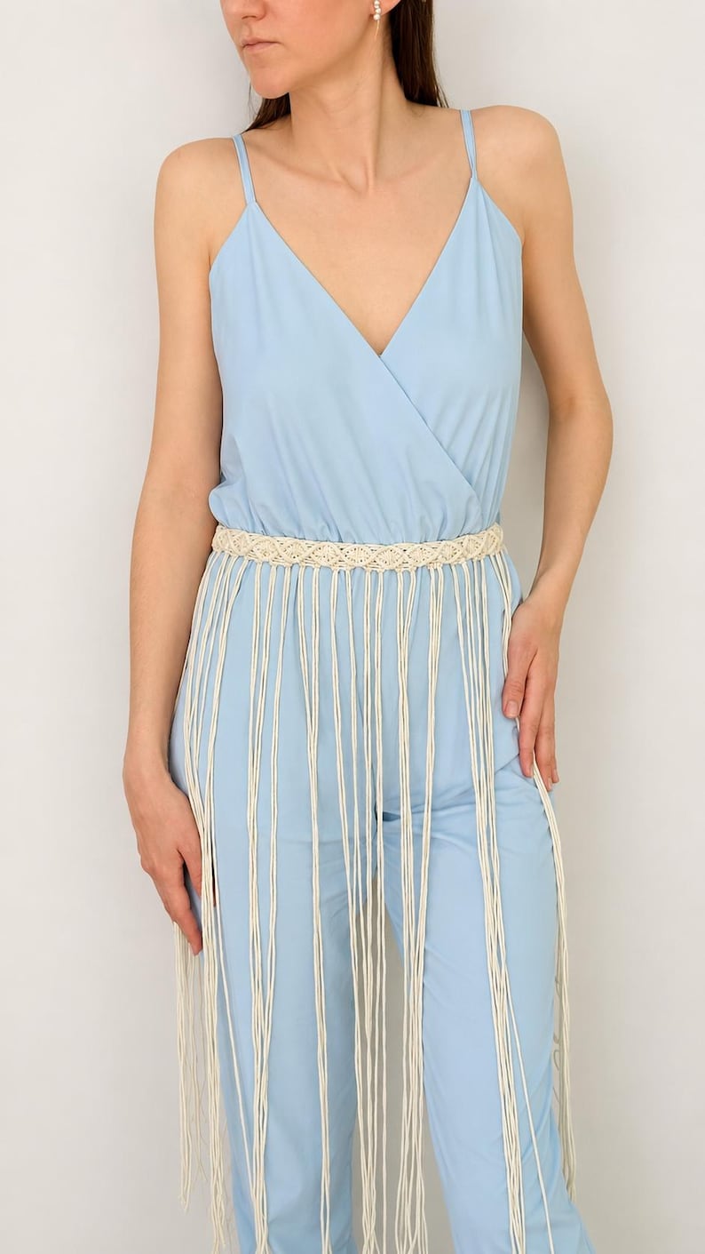 May include: A light blue sleeveless jumpsuit with a V-neckline and thin spaghetti straps, featuring a cream-colored macrame belt at the waist. The belt has a woven pattern and long, dangling fringe cords that extend down the front of the jumpsuit.