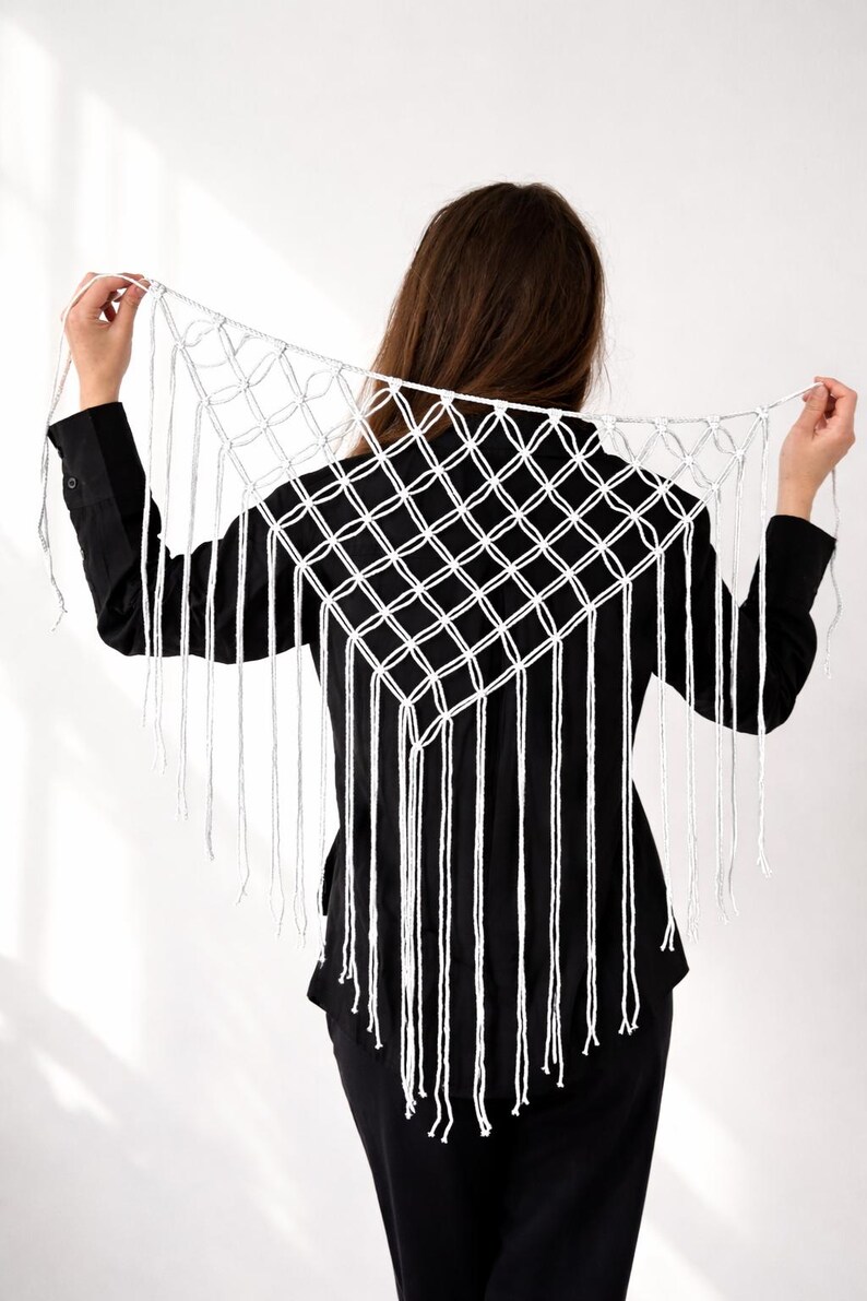 May include: A white macrame shawl with a geometric diamond pattern and long fringe. The shawl is held up against a black shirt, showcasing the intricate knotting and texture of the handmade piece. The design is a bohemian style.