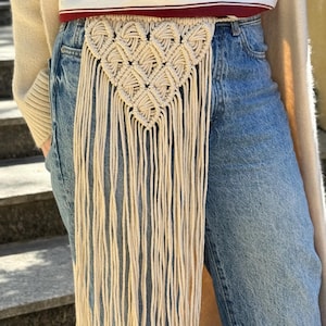 May include: A beige macrame belt featuring a triangular woven pattern with long, dangling fringe cords. The belt is worn over blue denim jeans, a white and maroon striped t-shirt, and a cream-colored open cardigan, creating a layered look.