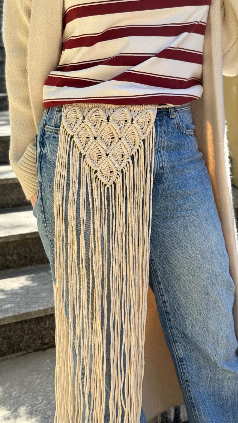 May include: A beige macrame belt featuring a triangular woven pattern with long, dangling fringe cords. The belt is worn over blue denim jeans, a white and maroon striped t-shirt, and a cream-colored open cardigan, creating a layered look.