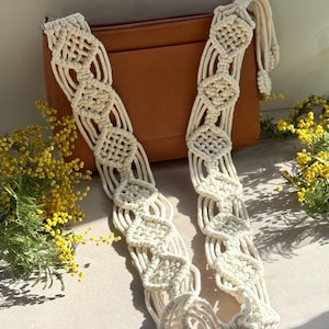 Puede incluir: Correa de bolso de macramé color crema con un diseño geométrico. La correa está colocada sobre un bolso de cuero marrón. Flores amarillas y follaje verde en el fondo.