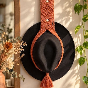 Può includere: Un cappello nero a tesa larga sostenuto da un supporto da parete in macramè color arancio bruciato. Il supporto presenta intricati motivi a nodi, due piccole perline di legno bianco, un bastoncino di legno e una lunga nappa centrale.