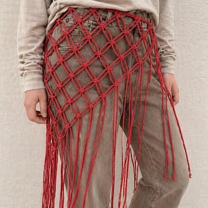 May include: A red macrame hip wrap featuring a knotted diamond grid pattern and long, dangling fringe strands. The accessory is worn over grey denim jeans and a long-sleeved beige shirt, creating a layered look with a textured, knotted cord design.