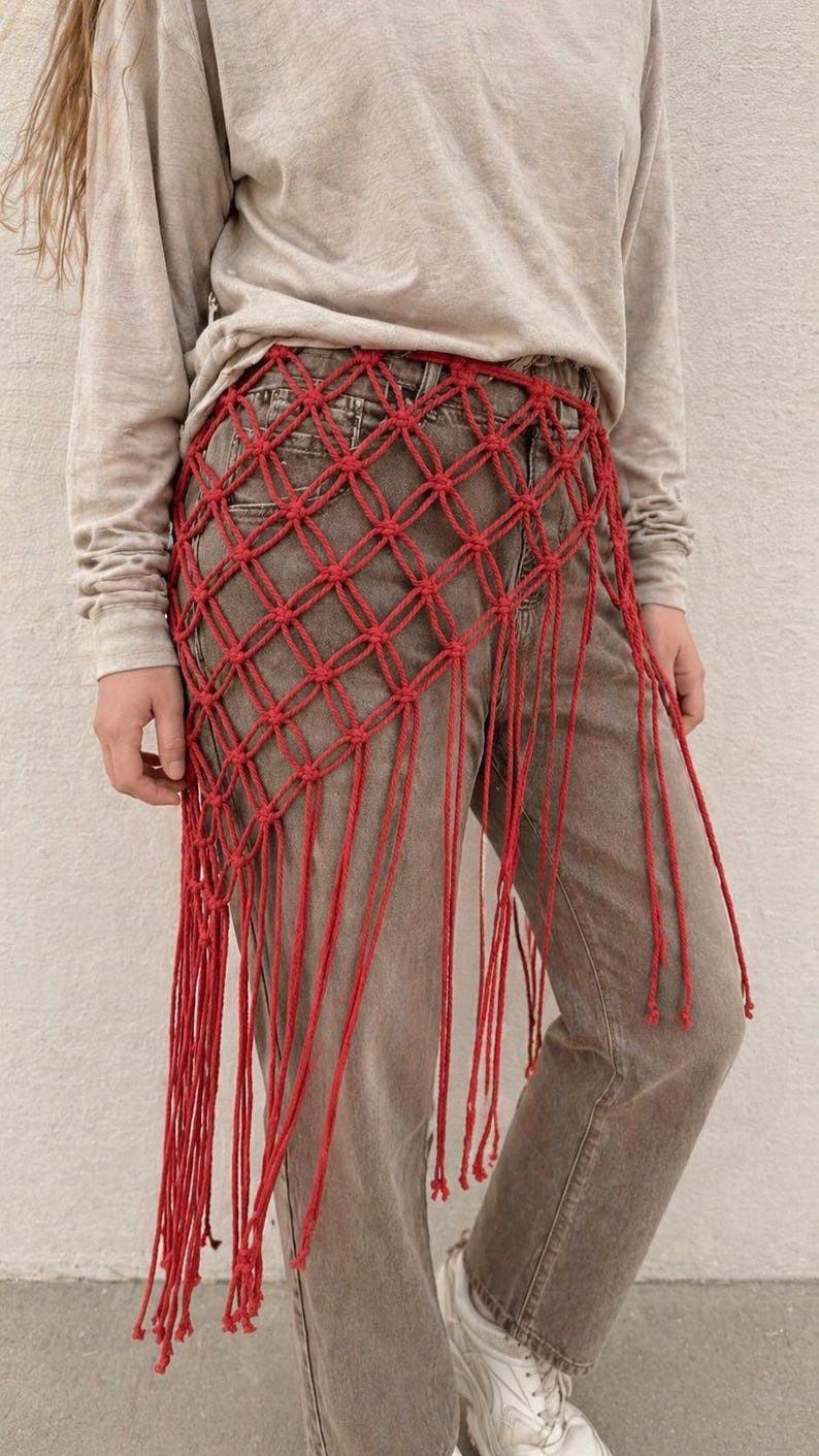 May include: A red macrame hip wrap featuring a knotted diamond grid pattern and long, dangling fringe strands. The accessory is worn over grey denim jeans and a long-sleeved beige shirt, creating a layered look with a textured, knotted cord design.