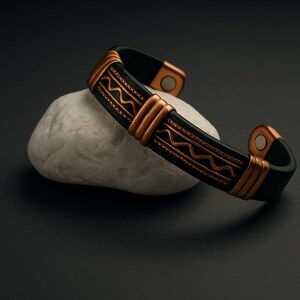 May include: A black cuff bracelet with copper-colored accents. The bracelet features a geometric design and is resting on a white stone against a dark background. The copper accents are on the edges and in the center of the bracelet.