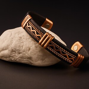 May include: A black leather bracelet with copper-colored metal accents. The bracelet features a decorative wave pattern and is resting on a white stone against a dark background. The bracelet has a magnetic clasp.