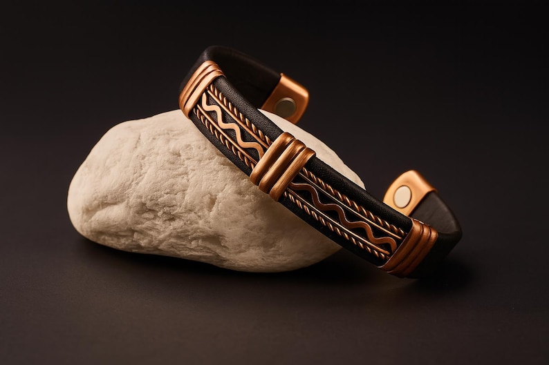May include: A black leather bracelet with copper-colored metal accents. The bracelet features a decorative wave pattern and is resting on a white stone against a dark background. The bracelet has a magnetic clasp.