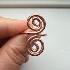 May include: Copper wire ring featuring two spiral designs. The ring is a warm copper color and has a minimalist, artistic style. The spirals are symmetrical and create a unique, eye-catching design. This handcrafted ring is a piece of jewelry.
