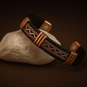 May include: A black leather cuff bracelet with copper-colored metal accents. The bracelet features a geometric design and is resting on a white stone. The metal has a decorative wave pattern.