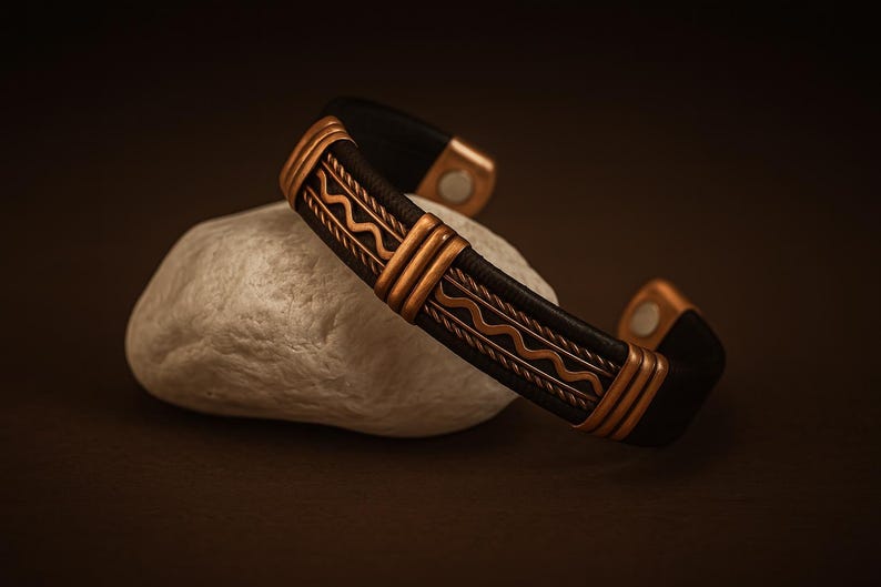 May include: A black leather cuff bracelet with copper-colored metal accents. The bracelet features a geometric design and is resting on a white stone. The metal has a decorative wave pattern.