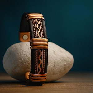 May include: A black leather bracelet with gold-colored metal accents and a magnetic clasp. The bracelet features a decorative design with wavy and linear patterns. The bracelet is displayed on a white stone against a dark teal background.