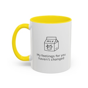 May include: White ceramic mug with a yellow interior and handle. The mug features a black and white illustration of a milk carton and the text "My feelings for you haven't changed."
